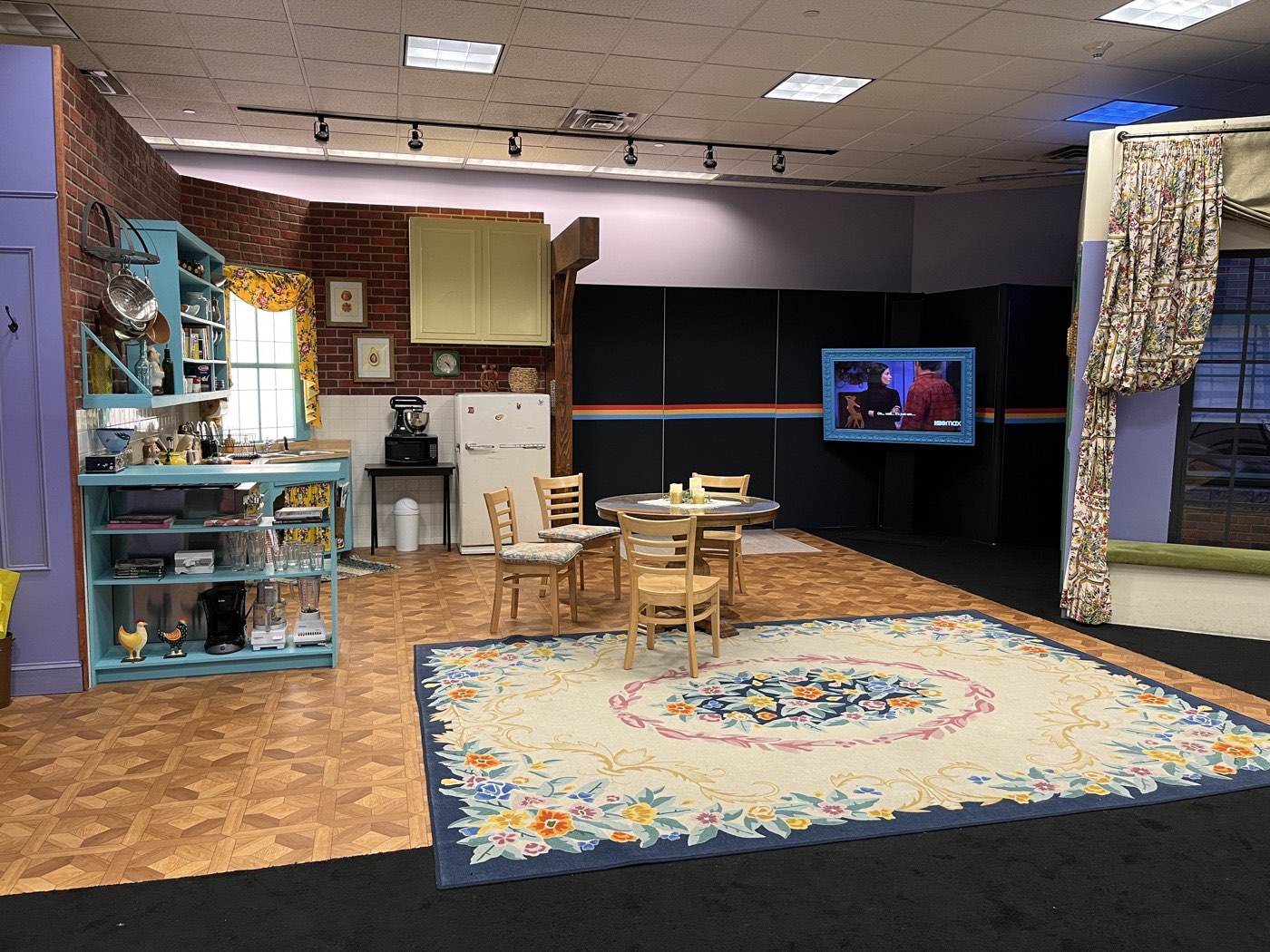 Recreation of Monica's apartment set: the turquoise kitchen cabinets, the small dining table with mismatched chairs in the middle of the open space, and the big rug where the living area begins.