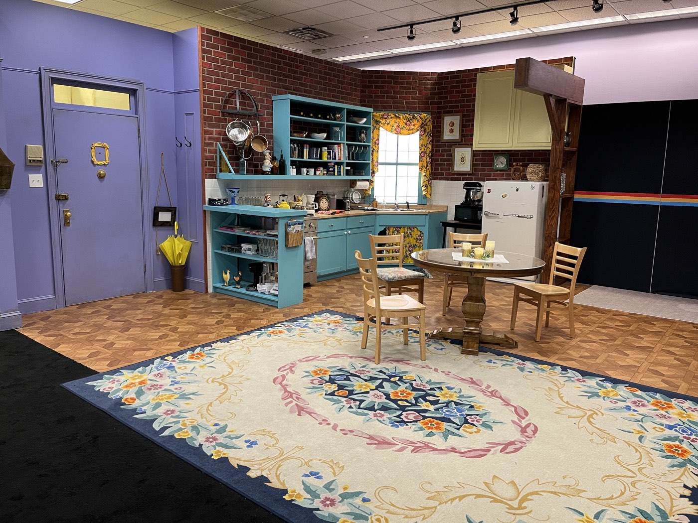 The kitchen corner of the Monica's apartment set recreation, with the small round-backed dining table and chairs parked between the kitchen cabinets and the open living space.